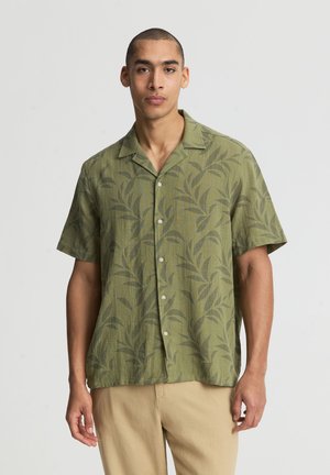 Giovane uomo in piedi che indossa una camicia verde oliva a maniche corte con bottoni e motivi a foglie e pantaloni beige su uno sfondo chiaro.