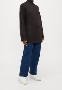 Person, der einen dunkelbraunen Rollkragenpullover, weit geschnittene blaue Jeans und weiße Sneakers trägt und vor einem einfarbigen Hintergrund steht.