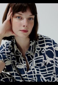 Jeune femme aux cheveux bruns courts et aux yeux bleus, appuyant sa tête sur sa main, portant une chemise à motifs bleu marine et blanc boutonnée.