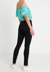 Teal off-the-shoulder kukkakuvioinen pusero pitsireunalla, yhdistettynä mustiin kapeisiin farkkuihin ja valkoisiin liukusandaaleihin, nähtynä takaa.