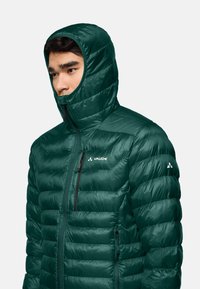Giacca a vento verde isolata con cappuccio, design trapuntato, zip completa e tasca laterale con zip. Presenta il logo sul petto e sulla spalla.