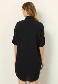 Rochie neagră din cămașă cu guler, mâneci scurte cu răsucire și croială dreaptă. Prezintă o textură netedă și un tiv curbat în partea de jos.