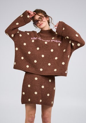 Kvinde iført brun, oversized polkaprikket trøje og matchende nederdel, justerer briller, neutral baggrund.