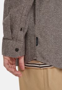Bruine, gestructureerde langetijdshirt met knoopmanchetten, met een merketiket aan de zijkant en een gestreept patroon dat eronder zichtbaar is.