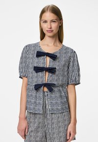 Top a righe in blu navy e bianco, con maniche a sbuffo corte, scollo rotondo e tre decorazioni a fiocco sul davanti. Materiale in cotone leggero.