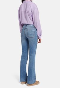 Camisa de manga larga de color púrpura claro, jeans de tiro alto acampanados de color azul con un acabado desvanecido y dobladillo sin terminar, combinados con calzado de color beige claro.