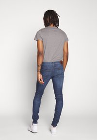 Camiseta gris de manga corta combinada con jeans ajustados azules. Los jeans tienen una etiqueta de cuero en la parte trasera, acompañados de zapatillas blancas.