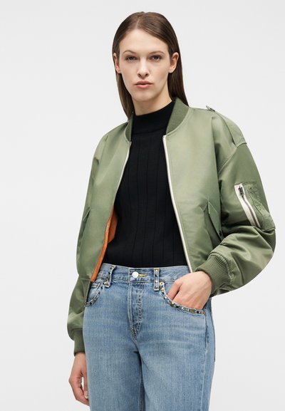 Γυναίκα με ίσια καστανά μαλλιά που φορεί μία μπουφάν bomber χακί πάνω από ένα μαύρο ριμπ πλεκτό πουλόβερ και ανοιχτό μπλε ψηλόμεσα τζιν με τσέπες διακοσμημένες με καρφιά.