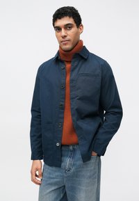 Tengerészkék gombos dzseki mellzsebbel, narancssárga garbóval párosítva. A modell világoskék farmert visel. Simat textúra.