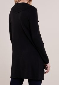 Lång svart cardigan i ribbat material. Har en bred krage och sidokilar för rörelse. Snygg, enfärgad design.