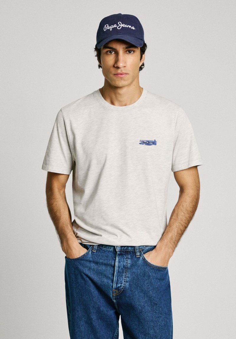 Light gray cotton t-shirt with a round neck and blue graphic detail on the chest, paired with blue denim jeans and a navy cap.