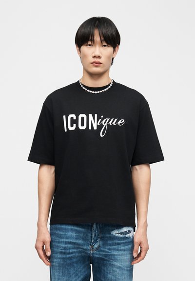 Νεαρός με ίσια μαύρα μαλλιά φοράει μαύρο μπλουζάκι με την επιγραφή "ICONique" και κολιέ, συνδυασμένα με ξεφτισμένο μπλε τζιν.