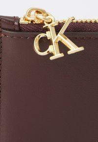 Rudas odinis maišelis su auksiniu užtrauktuku ir auksiniu „cK“ logotipo žavesiu. Matoma lygi tekstūra ir tvarkingi siuvimo detalės.