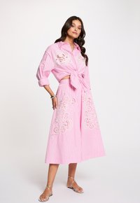 Mujer vestida con un conjunto de dos piezas rosa con rayas y bordado floral con recortes, camisa anudada, falda de pierna ancha y sandalias plateadas.