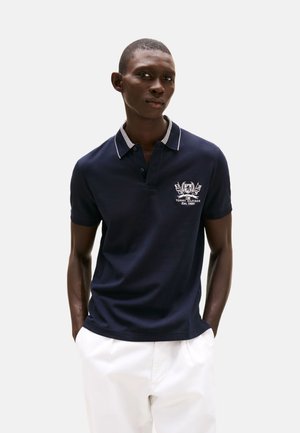 Camisa polo azul marino con cuello a rayas, mangas cortas y logo bordado. Combinada con pantalones blancos, que presentan una textura de tela suave.