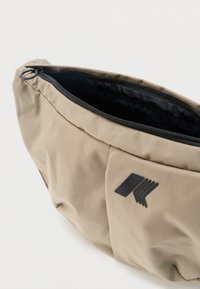 Μπεζ τσαντάκι μέσης από ύφασμα με μαύρο φερμουάρ που είναι μερικώς ανοιχτό, αποκαλύπτοντας την μαύρη εσωτερική επένδυση και ένα μικρό μαύρο γεωμετρικό λογότυπο στο μπροστινό μέρος.