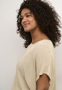 Beige, loose-fitting top featuring a textured fabric, round neckline, and short sleeves, displayed on a model with curly hair.