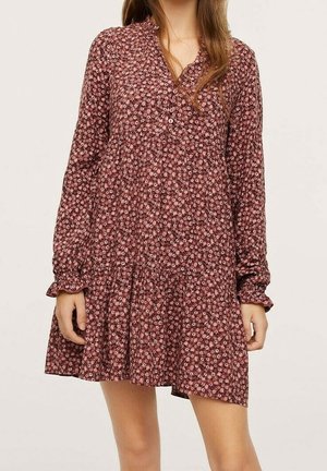 Kvinde iført en langærmet, knappet, løstsiddende kjole med et rødt og pink blomsterprint, stående mod en ensfarvet baggrund.