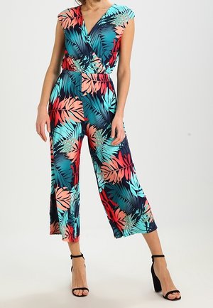 Femme portant une combinaison sans manches à imprimé feuilles tropicales multicolores, avec un pantalon large et court, et des sandales à talons hauts noires.