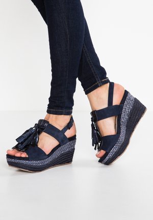 Sandales compensées bleu marine avec brides à la cheville et détails à pompons, portées avec un jean skinny foncé sur un fond blanc net.