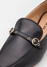 Mocassin en cuir noir avec détail boucle dorée et bride sur le dessus, finition lisse texturée et doublure intérieure beige.