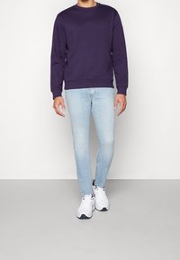Sudadera de cuello redondo morada de manga larga, combinada con jeans ajustados azul claro y zapatillas blancas, de pie contra un fondo simple.