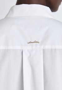 Camicia bianca con un design plissettato sul retro e un piccolo accento in oro vicino al colletto. Tessuto liscio e leggero.