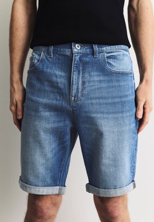 Man draagt blauwe opgerolde denimshorts en een zwart shirt, staat met ontspannen handen langs zijn zij tegen een effen achtergrond.