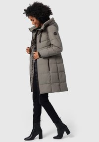 Gevoerde, knielange jas in gedempt grijs, met een capuchon, geborduurd textuur, bruine accenten en ritszakken aan de zijkanten. Draagt over zwarte broeken en boots.