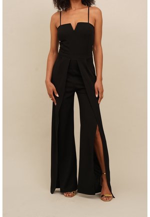 Femme portant une combinaison noire sans manches avec des fentes à l'avant sur un pantalon à jambes larges et des sandales ouvertes dorées sur un fond beige.