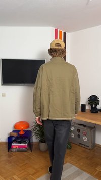 Χακί φαρδύ μπουφάν, μαλακό ύφασμα, ψηλός γιακάς, αντίθεση ραφής, συνδυασμένο με σκούρα παντελόνια. Το φόντο περιλαμβάνει μια τηλεόραση και πολύχρωμα διακοσμητικά.