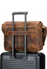 Una borsa a tracolla in pelle marrone con pattina è posata su una valigia nera con ruote. Presenta una tasca con zip e una tracolla.
