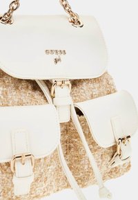 Biały plecak z sztucznej skóry z plecionymi brązowymi akcentami, złotym metalowym wykończeniem i zapięciem na magnes. Posiada dwie przednie kieszenie z klamrami.