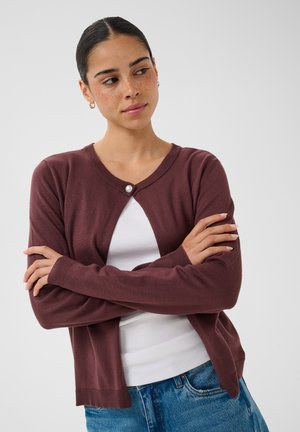 Jeune femme aux cheveux foncés et taches de rousseur portant un cardigan bordeaux par-dessus une chemise blanche et un jean bleu, bras croisés, regardant sur le côté.