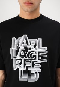 Fekete pamut póló, amelyen előtérbe helyezett fehér 3D szöveggrafika található, a "KARL LAGERFELD" felirattal, rétegezett és árnyékos betűtípussal.