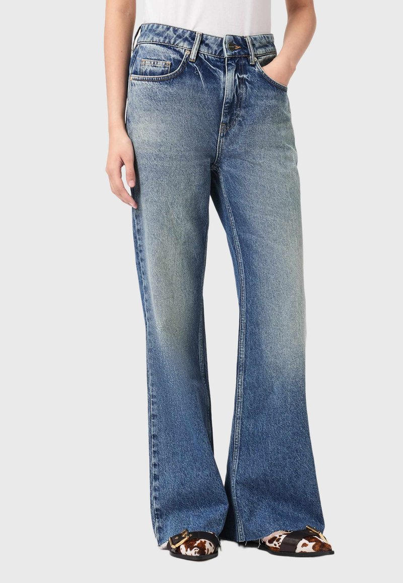 Jeans mit hohem Bund, blauem Denim und ausgestelltem Bein sowie einem hellen Wasch-Effekt, ausgestattet mit klassischen Taschen und Reißverschluss, kombiniert mit gemusterten flachen Schuhen.