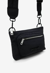 Black nylon crossbody bag with front zip pocket, embossed logo, adjustable strap, and silver-tone hardware. Smooth texture, rectangular shape.