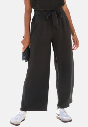 Person trägt hoch taillierte schwarze weite Hose mit gebundener Gürtel, weiße Turnschuhe, hält eine dunkel strukturierte Clutch-Tasche.