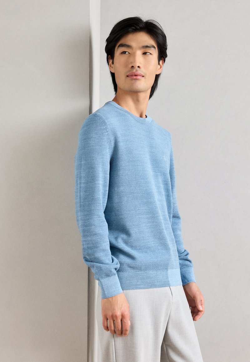 Ανοιχτό μπλε πλέκτο πουλόβερ με στρογγυλή λαιμόκοψη και ριγέ μανσέτες. Λείος υφασμάτινος χαρακτήρας και διακριτό σχέδιο σε όλη την επιφάνεια. Συνδυασμένο με ανοιχτό γκρι παντελόνι.