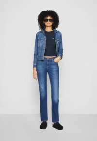 Giacca di denim, top corto blu scuro con logo, jeans azzurri a gamba dritta, scarpe nere slip-on, occhiali da sole, capelli ricci.
