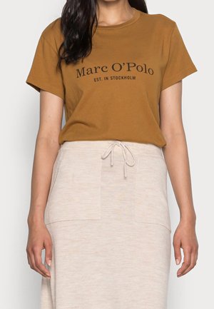 Camiseta de algodón marrón con mangas cortas, que presenta el texto negro "Marc O’Polo EST. IN STOCKHOLM." Combinada con una falda beige clara con bolsillos frontales.