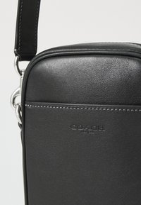 Borsa a tracolla nera in pelle con texture liscia, bordi arrotondati, hardware in argento e logo impresso. Caratterizzata da una tracolla piatta per indossarla.