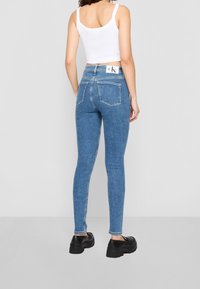 Jeans de denim azul ajustados con una cintura alta, cinco bolsillos y un parche de logo blanco en la parte trasera. Tejido texturizado con un acabado suave.