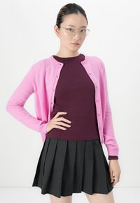 Vrouw met een bril die een roze cardigan draagt over een bordeauxrode top en een donkere geplooide rok, poserend met één hand op haar heup.