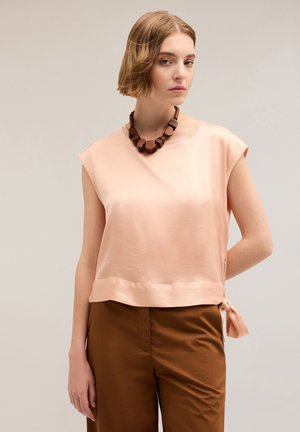 Top senza maniche di un leggero color pesco con scollatura ampia, dettaglio di nodo laterale, abbinato a pantaloni marroni. Tessuto liscio, design casual.