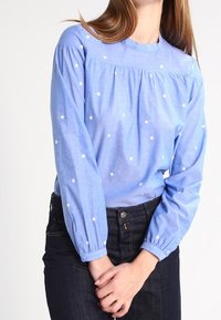 Blouse bleu clair à manches longues, avec broderie d'étoiles blanches, encolure froncée et poignets élastiqués. Associée à un jean foncé.