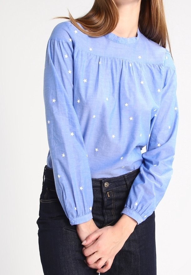 Blouse bleu clair à manches longues, avec broderie d'étoiles blanches, encolure froncée et poignets élastiqués. Associée à un jean foncé.