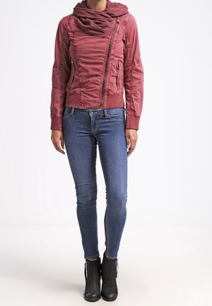 Kvinde iført en støvet rosa asymmetrisk lynlåstrøje, blå skinny jeans, sorte ankelstøvler, stående mod en hvid baggrund.