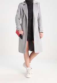 Cappotto grigio chiaro di media lunghezza con revers a punta, texture morbida; vestito nero a collo alto sotto, abbinato a sneaker bianche e una pochette.