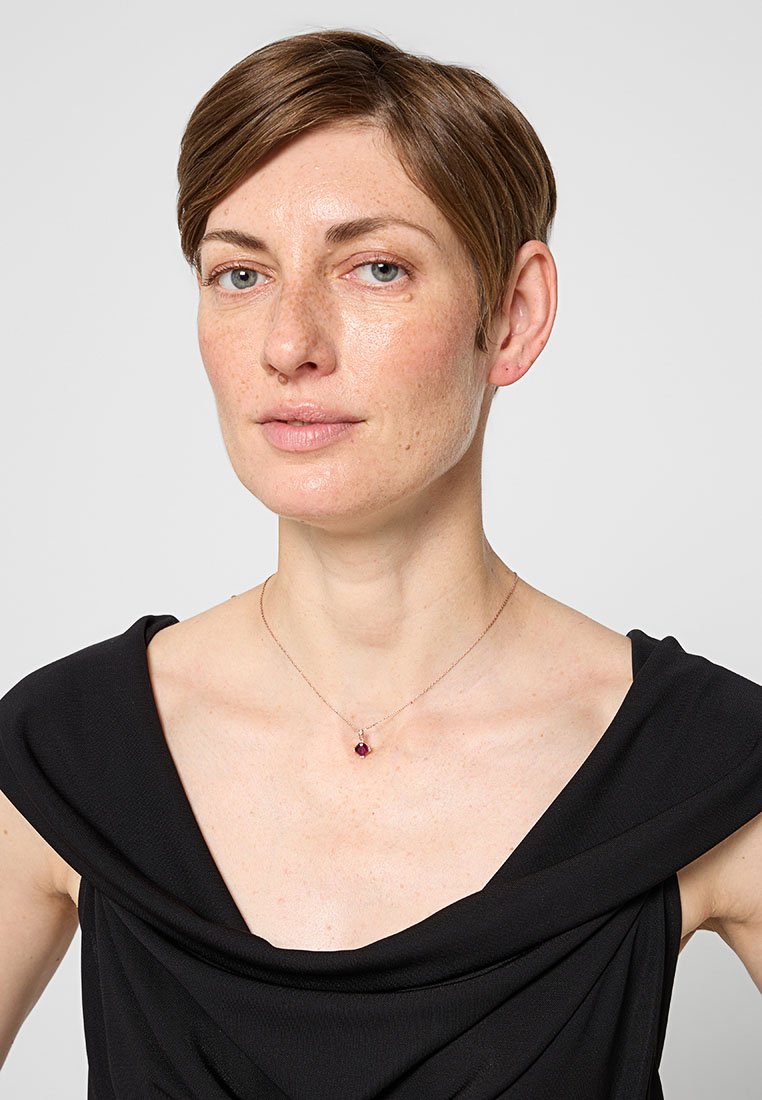 Collana d'oro con una piccola pietra preziosa rotonda viola, indossata su un collo nudo, contro un top nero drappeggiato. La pietra preziosa riflette la luce in modo sottile.